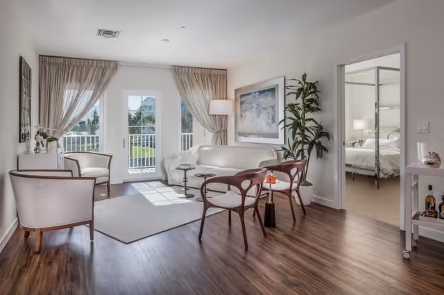 A bright and airy living room with wooden flooring, a white sofa, two wooden chairs with white cushions, a small round side table, a large potted plant, and a floor lamp. French doors with beige curtains open to an outdoor area. A doorway on the right leads to a bedroom with a bed and bedside lamp visible.
