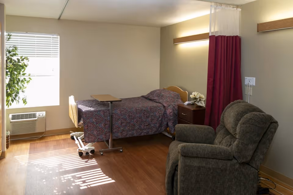 A small, simple bedroom in a senior living facility with a single bed covered in a patterned bedspread, a bedside table with a flower arrangement, a reclining chair, a window with blinds, and a portable overbed table. The room has wooden flooring and neutral-colored walls with soft lighting.