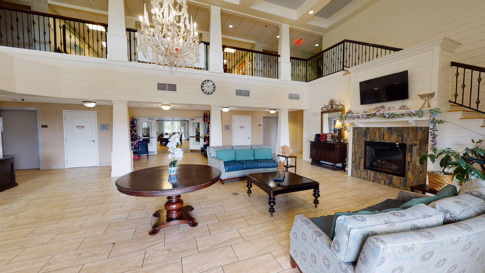 Spacious senior living facility common area with high ceilings, a large chandelier, and a balcony railing overlooking the room. The room features a round wooden table with a flower vase, two sofas with cushions, a coffee table, a fireplace with a TV mounted above it, and decorative plants. The floor is tiled, and there are multiple doors and a clock on the far wall.