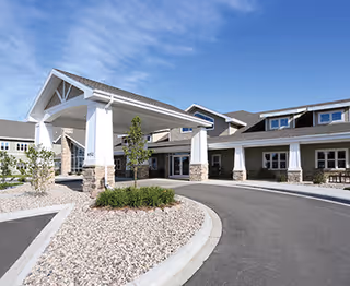 Exterior view of a senior living facility named Homestead I and Homestead II, showing a covered entrance with white pillars and stone bases, a curved driveway, landscaped areas with rocks and small plants, and a clear blue sky.