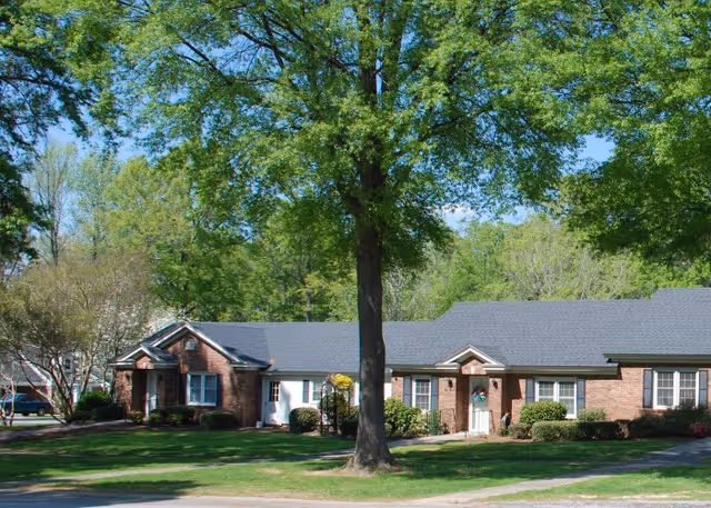 Exterior view of a single-story brick building with a gray roof, surrounded by green grass and trees under a clear blue sky.