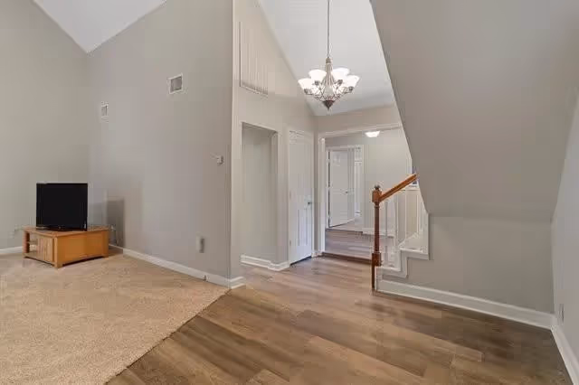 Interior view of a residential space with a small wooden TV stand and television on the left, beige carpet flooring in the living area, wood flooring in the hallway, a staircase with wooden handrails on the right, and a chandelier hanging from the ceiling.