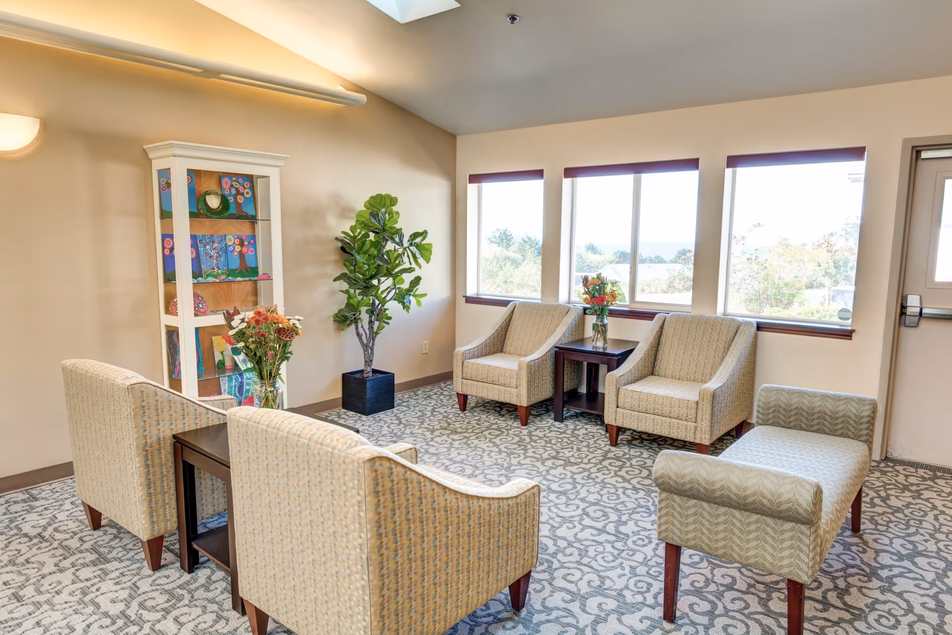 A bright and cozy sitting area with several upholstered chairs and a patterned carpet. There is a small table with a vase of flowers between two chairs near three large windows letting in natural light. A tall plant and a white display cabinet with colorful artwork are against the wall.