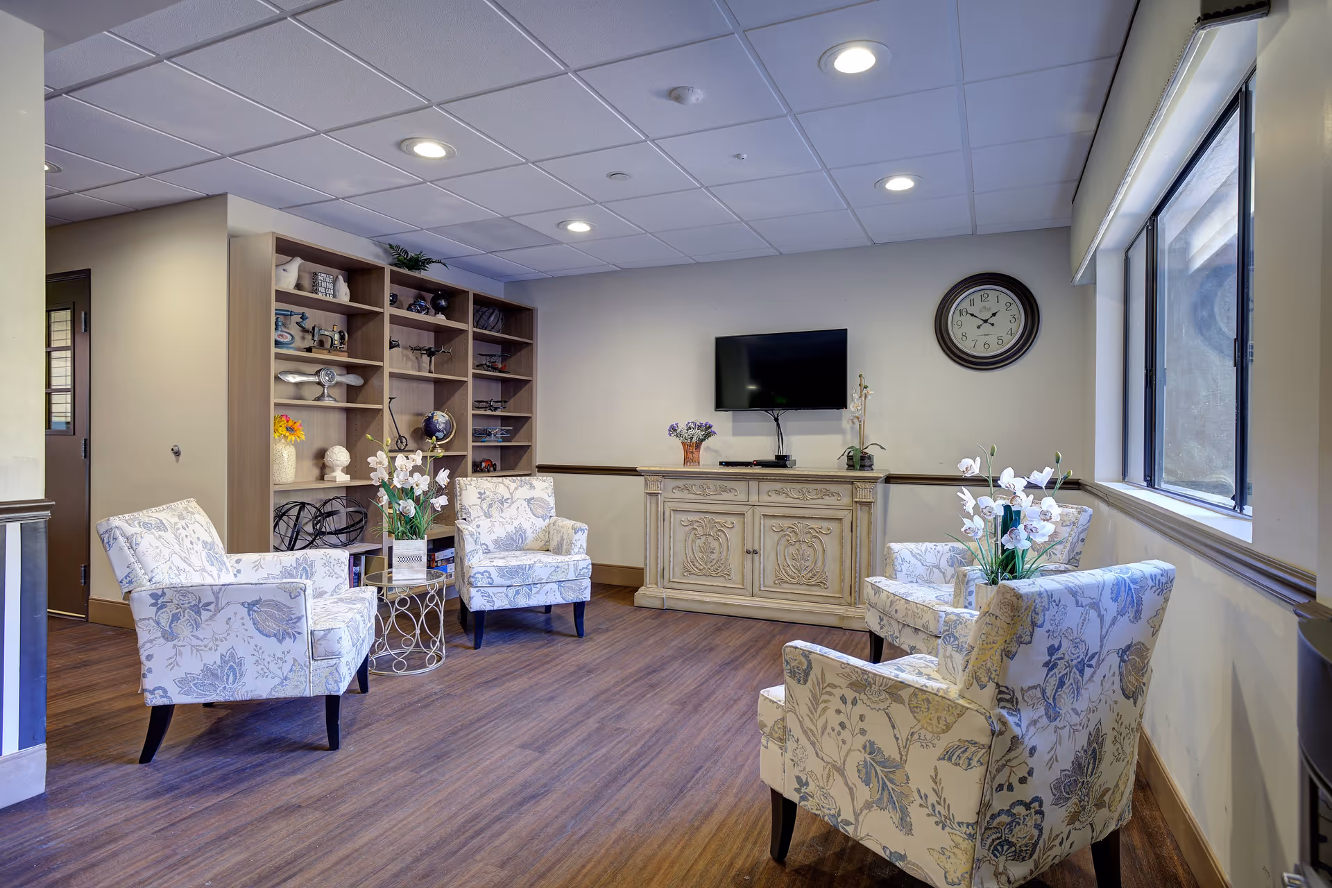 A cozy senior living facility common area with four floral-patterned armchairs arranged around a small round table with a flower vase. There is a wooden cabinet with a flat-screen TV mounted above it and a wall clock to the right. A large window lets in natural light, and a bookshelf filled with decorative items is visible in the background.