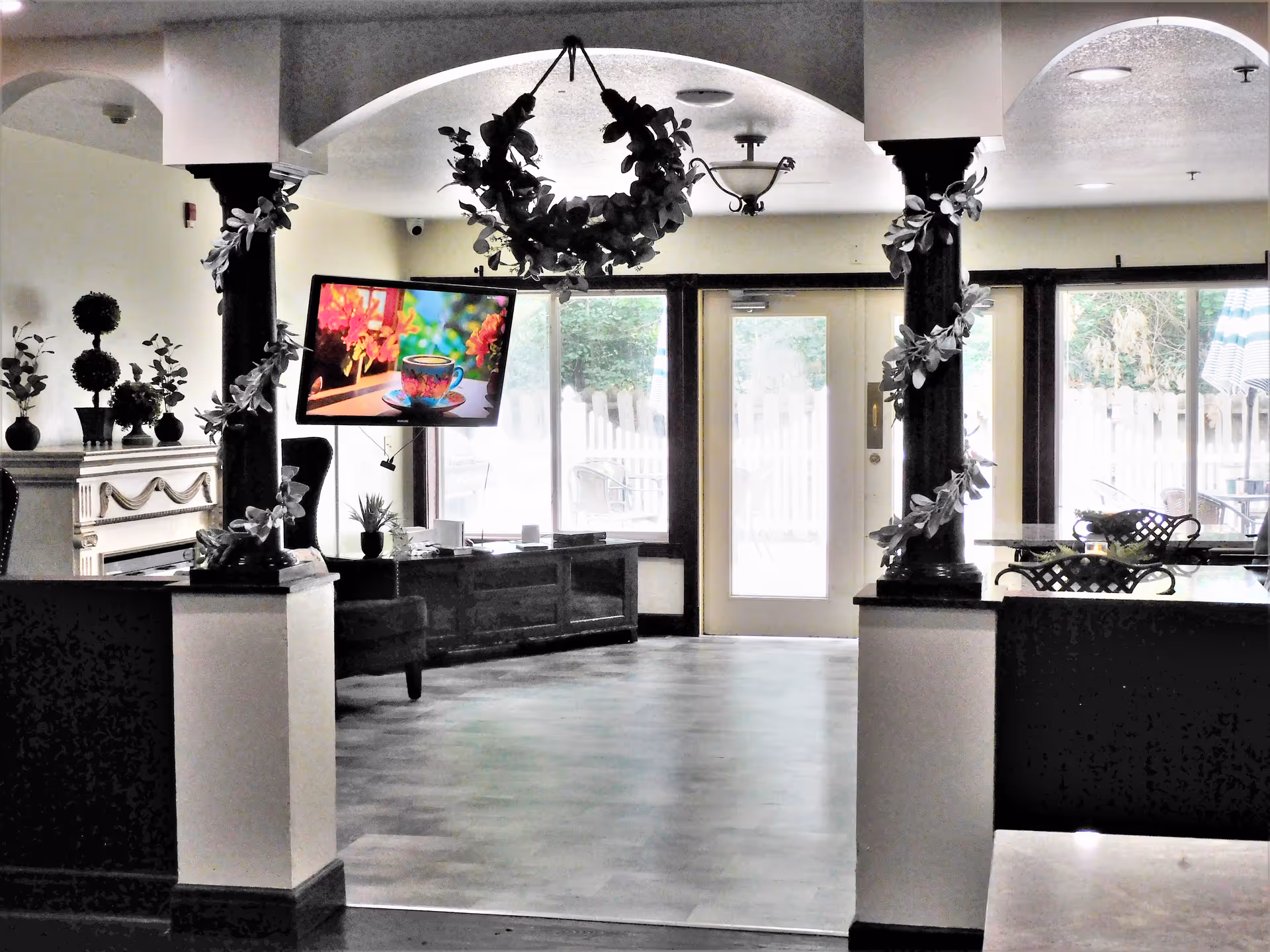 Interior view of a common area in Cascades Assisted Living featuring two dark wooden columns wrapped with leafy garlands, a mounted flat-screen TV displaying a colorful image of a teacup and flowers, a fireplace with decorative plants on the mantle, and large glass doors and windows letting in natural light with a view of an outdoor patio area.