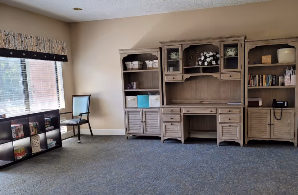 A quiet room with a large window on the left covered by blinds and a patterned valance. Below the window is a black shelving unit holding various board games and puzzles. Next to the shelving unit is a single wooden chair with a light blue cushion. Along the right wall, there are three wooden storage units with shelves, drawers, and cabinets. The middle unit has decorative items including a flower arrangement and small bird figurines. The room has beige walls and a blue carpeted floor.
