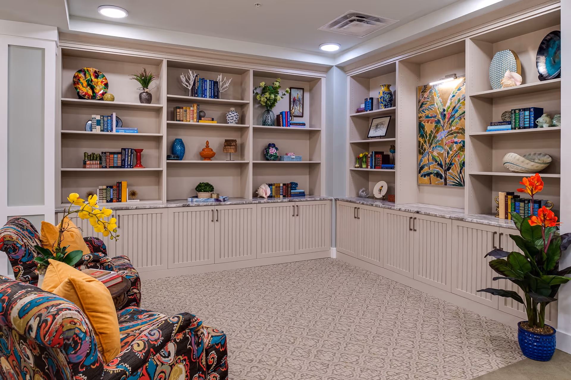 A cozy interior corner of a senior living facility featuring built-in beige bookshelves filled with books, decorative vases, and art pieces. Two colorful patterned armchairs with yellow cushions are positioned near a small round wooden table with books and a yellow orchid plant. The floor is carpeted with a subtle patterned design, and a large potted plant with red flowers is placed on the right side. The walls are light-colored, and the space is softly lit with ceiling lights.