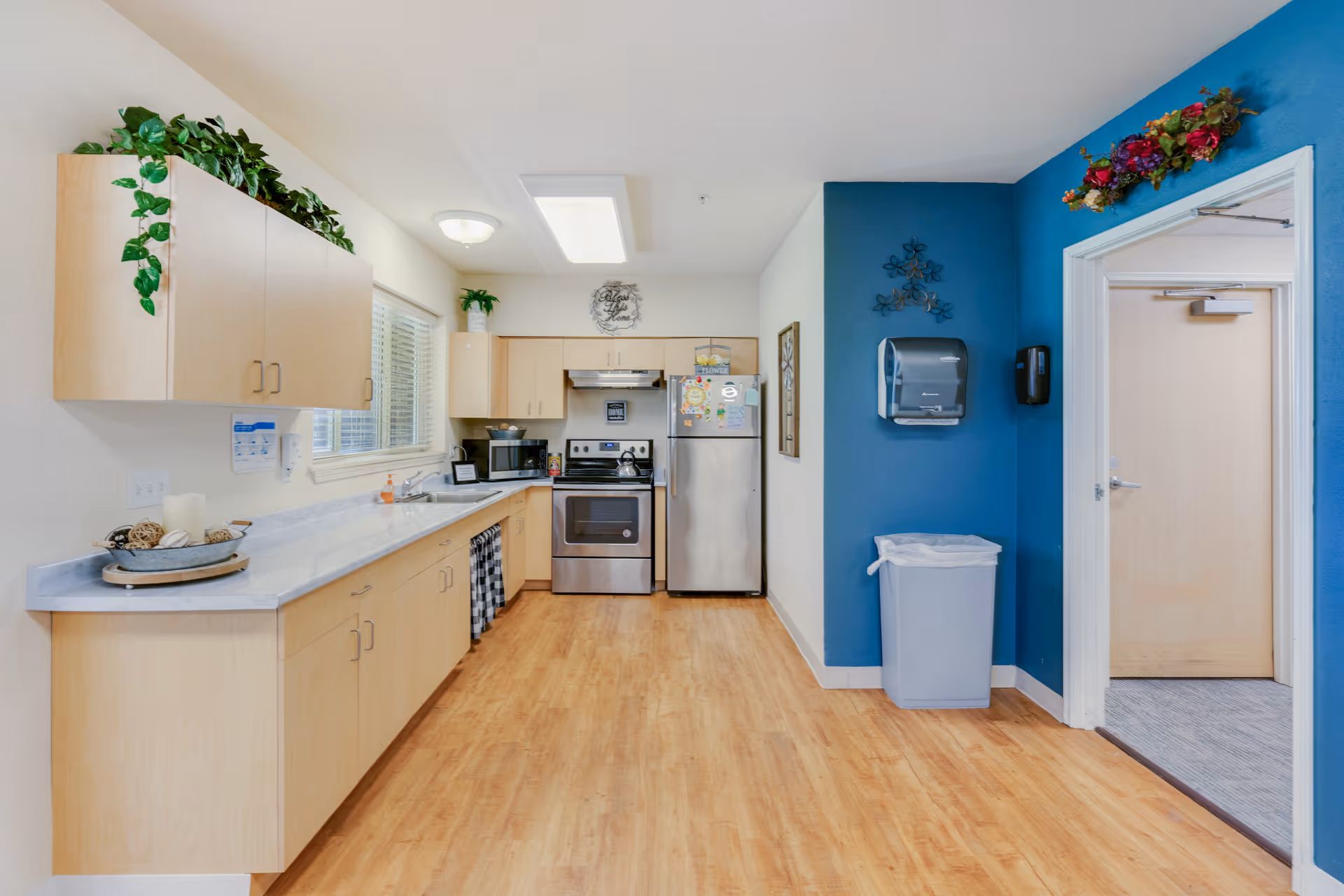 A bright kitchen area with light wood cabinets, a marble countertop, stainless steel stove and refrigerator, a microwave, and a window with blinds. The floor is wood, and one wall is painted blue with a trash bin and paper towel dispenser mounted on it. There are decorative plants on top of the cabinets and a floral arrangement above the doorway leading to another room.
