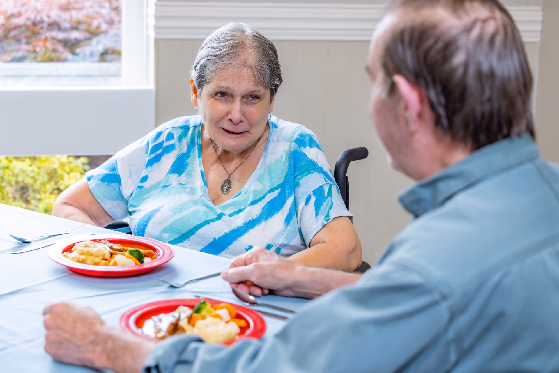 Two seniors sit at a dining table with plates of food, a woman facing the camera and a man seen from behind.