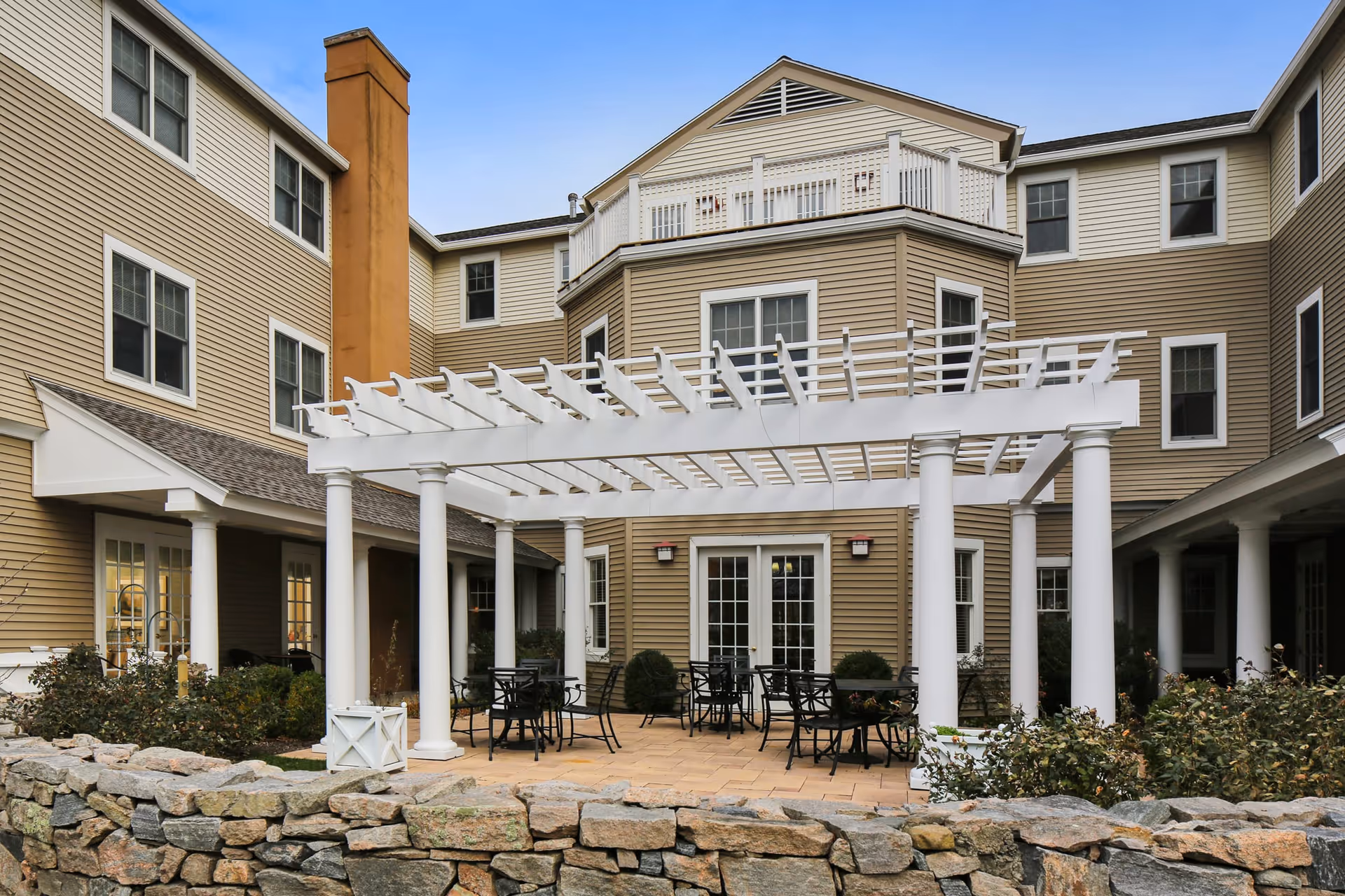 Outdoor courtyard area of a senior living facility with a white pergola, several black metal tables and chairs, surrounded by beige multi-story building with many windows and a stone wall in the foreground.