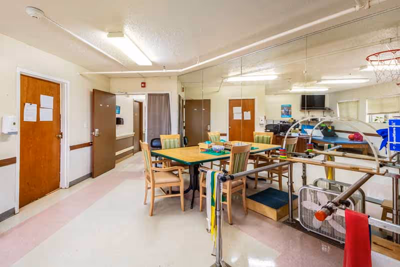 A rehabilitation room in a nursing facility with a table and chairs in the center, parallel bars for walking exercises, a basketball hoop mounted on the wall, a large mirror covering one wall, and doors leading to other rooms.