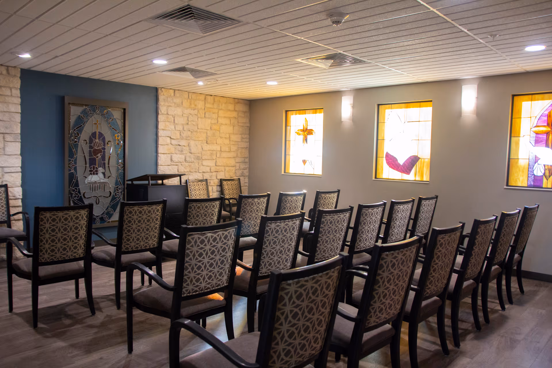 Chapel-style meeting room with rows of patterned chairs, a podium, and stained-glass windows on the wall.