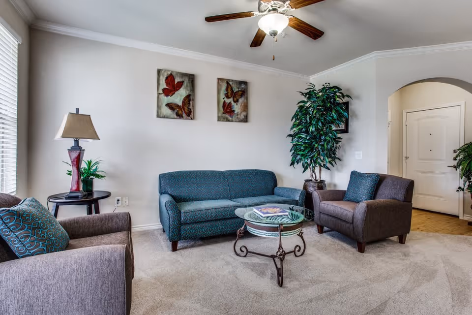 A cozy living room with a teal patterned sofa, two brown armchairs with teal patterned cushions, a glass coffee table with a book on it, a side table with a lamp and a small plant, two butterfly paintings on the wall, a large potted plant in the corner, and a ceiling fan with a light fixture.