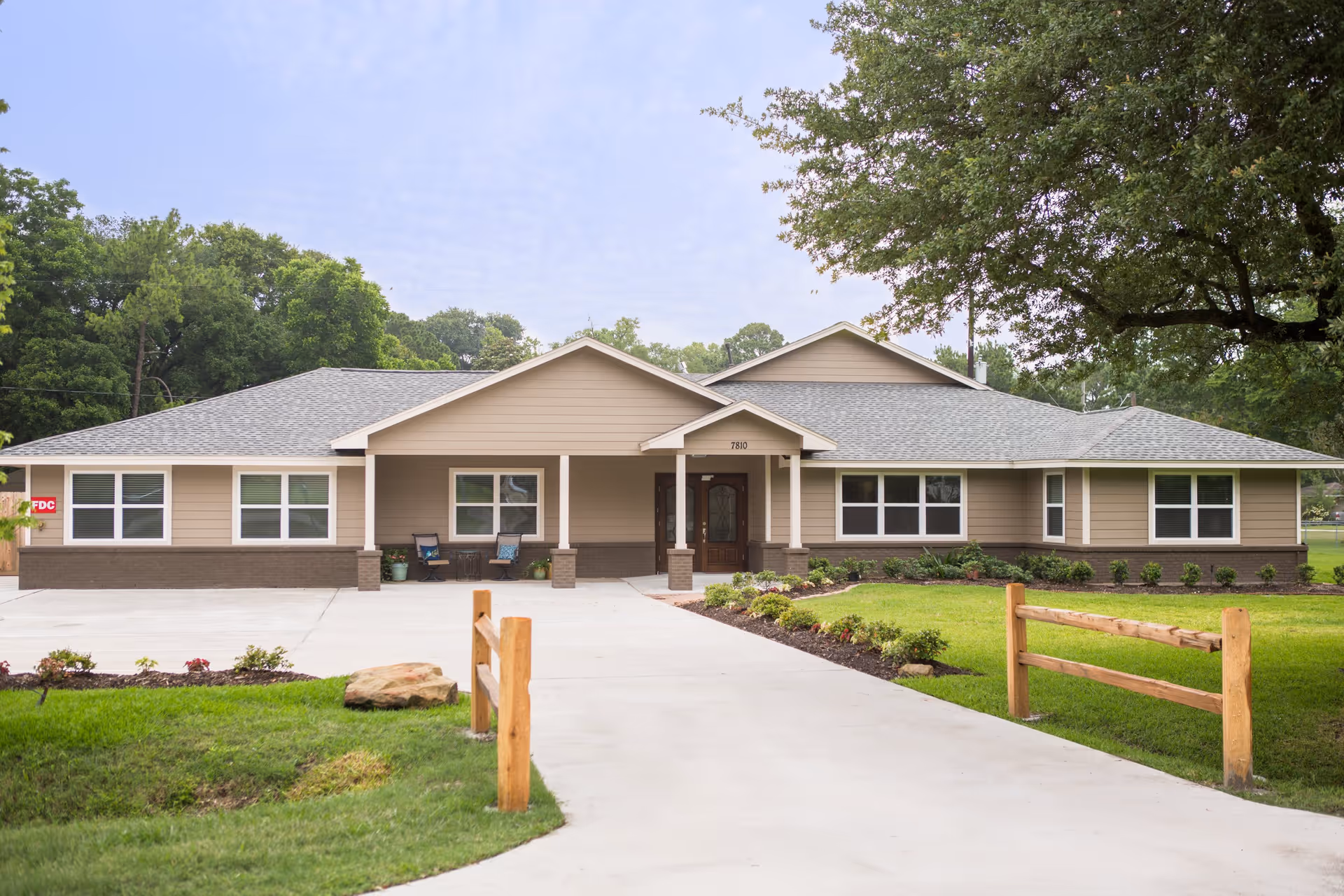 Front exterior view of a single-story assisted living facility building with beige siding and a gray shingled roof. The entrance has a covered porch with two chairs and potted plants. There is a concrete driveway leading up to the entrance, bordered by green grass, small shrubs, and wooden fencing. Large trees are visible in the background.