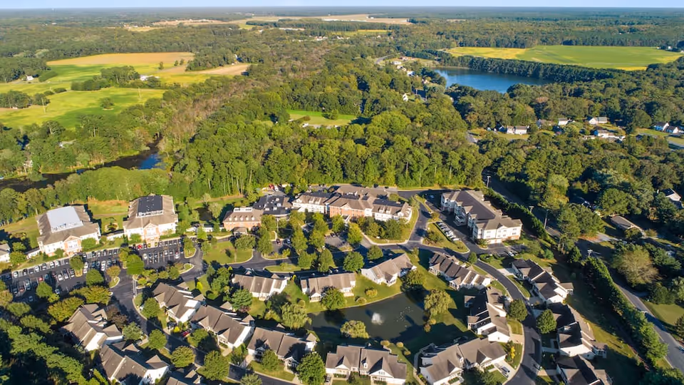Aerial view of Lakeside At Mallard Landing senior living facility surrounded by lush greenery, trees, and a small pond, with multiple residential buildings and parking areas visible under a clear sky.