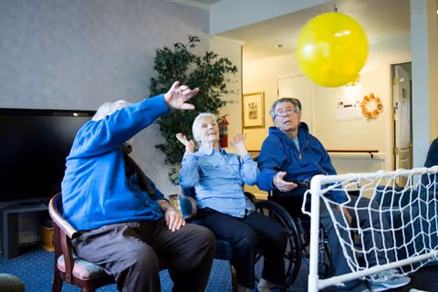 Three elderly individuals sitting in a room, engaging in a game with a large yellow balloon. One person is in a wheelchair, and a small goal net is visible in front of them. The setting appears to be a common area in a senior living facility.