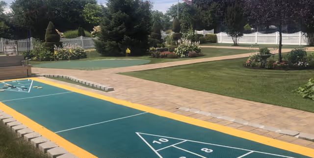 Outdoor garden area featuring shuffleboard courts, paved walkways, lawns, flower beds and a white picket fence.