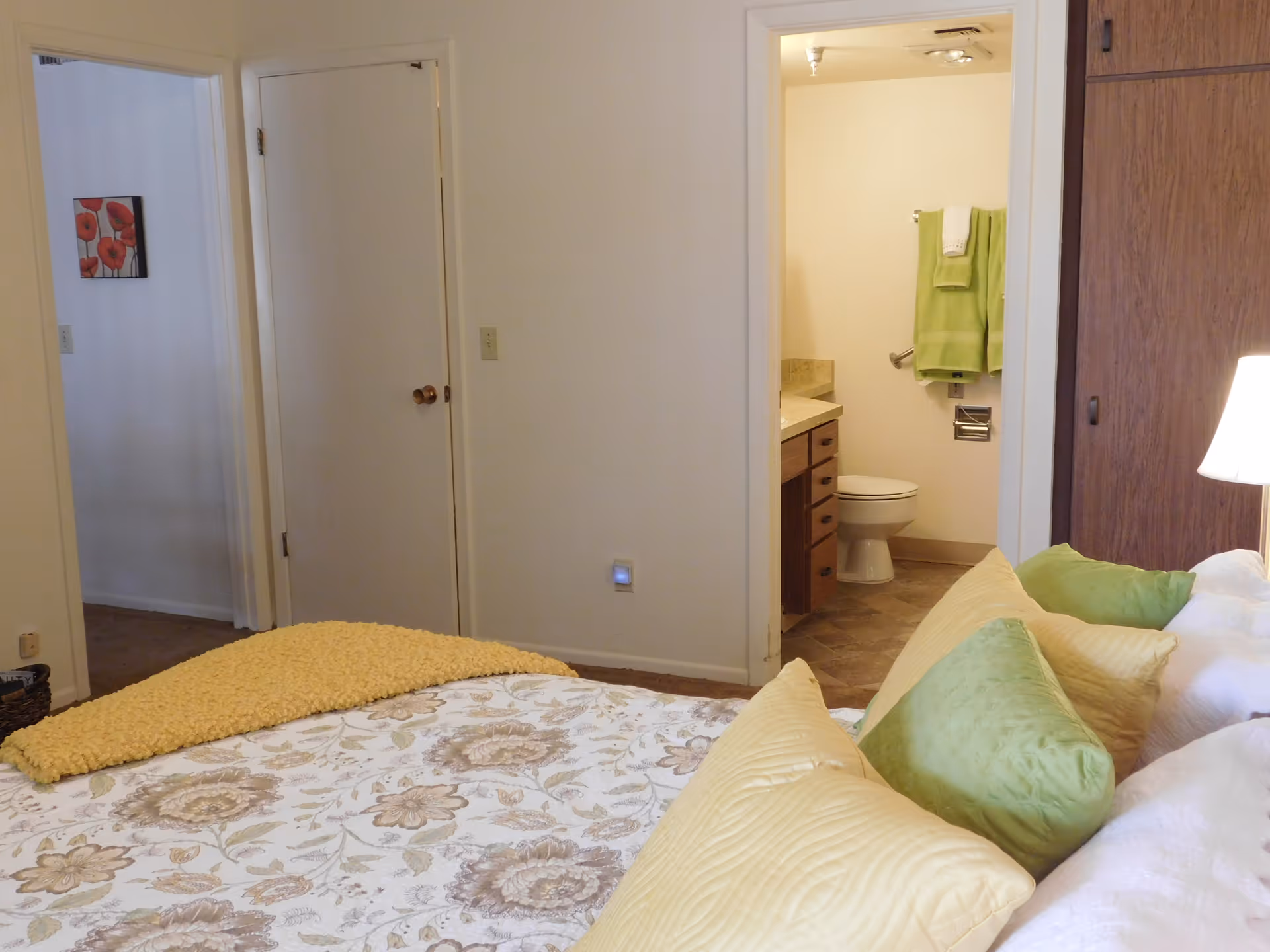 View of a bedroom with a bed covered in floral bedding and multiple pillows in shades of green and yellow. To the right, a bedside lamp is turned on. In the background, an open door reveals a bathroom with a toilet, countertop, and green towels hanging on the wall. Another door and a hallway with a framed picture of red flowers are visible on the left side.