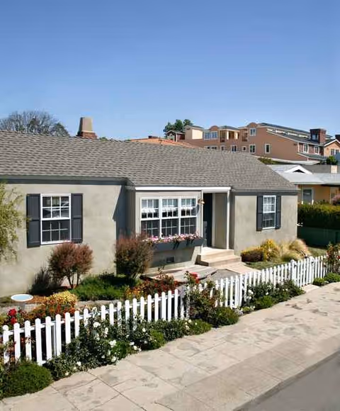 A single-story beige house with a white picket fence and colorful front garden.