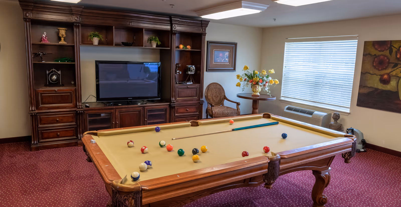 Carpeted interior game room with a billiards table, wooden entertainment center with a TV, chairs, and a window with blinds.