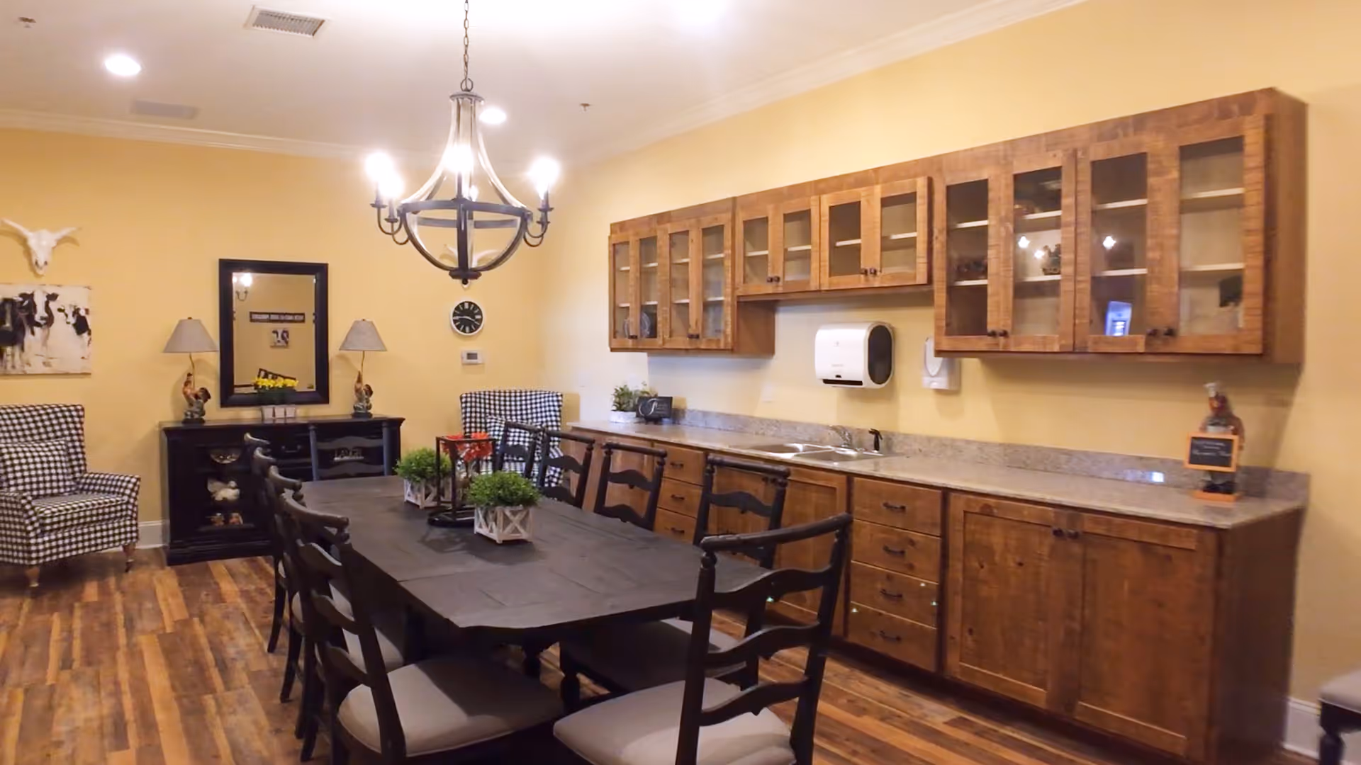 A dining room with a long dark wooden table surrounded by chairs. The room features wooden cabinets with glass doors mounted on the wall above a countertop with a sink. There are two checkered armchairs in the corner, a black sideboard with decorative items, a wall clock, and a chandelier hanging from the ceiling. The floor is wooden and the walls are painted yellow.