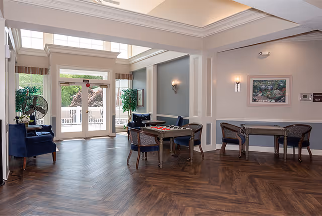 Bright communal lounge with card tables, upholstered chairs, and large glass doors leading to an outdoor patio.