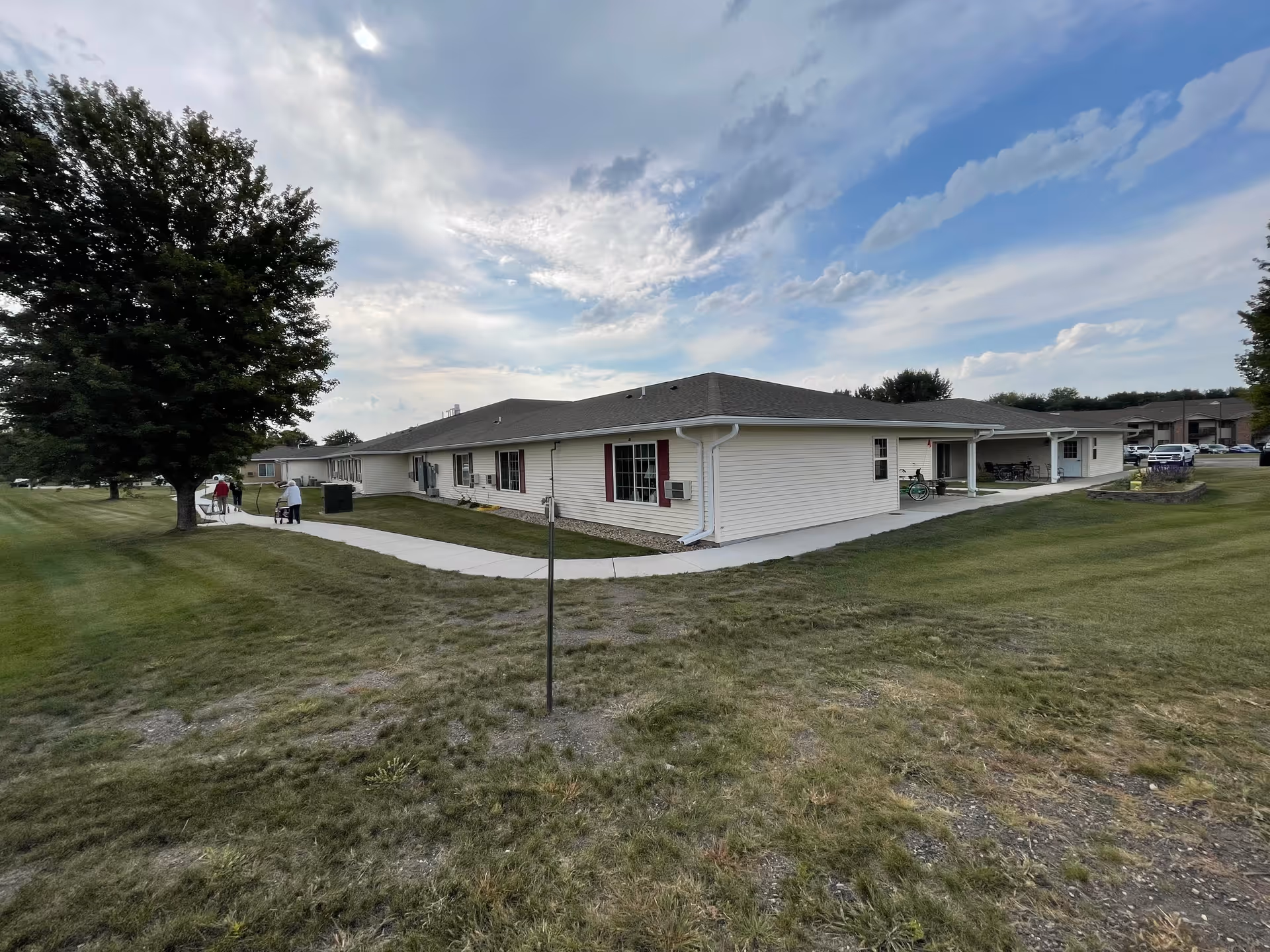 Exterior view of a single-story senior living facility building with white siding and a gray roof, surrounded by green grass and trees under a partly cloudy sky. A paved walkway runs alongside the building, and a few people are walking near the building.