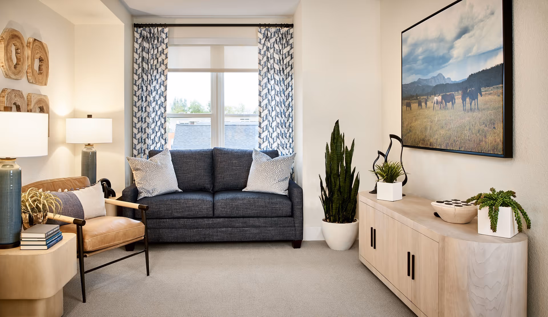A cozy living room with a dark blue sofa adorned with two light-colored pillows, a tan leather armchair with a pillow, and a light wooden side table with books and a plant. There are two matching lamps with blue bases on either side of the sofa. A large window with patterned curtains lets in natural light. On the right wall, there is a wooden cabinet with decorative plants and sculptures, and above it hangs a large framed picture of horses in a field with mountains in the background.