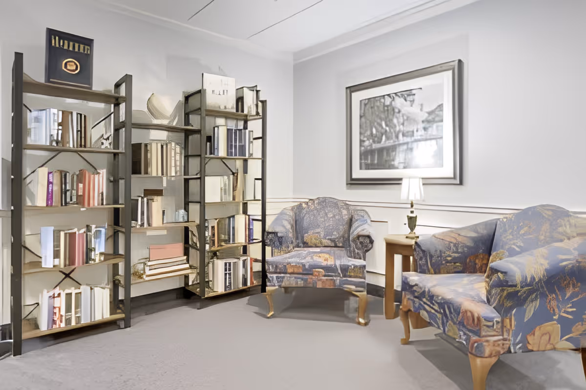 A cozy sitting area with two patterned armchairs and a small wooden side table with a lamp. Behind the chairs is a large framed black and white picture on the wall. To the left, there is a unique bookshelf with mirrored panels reflecting the room.