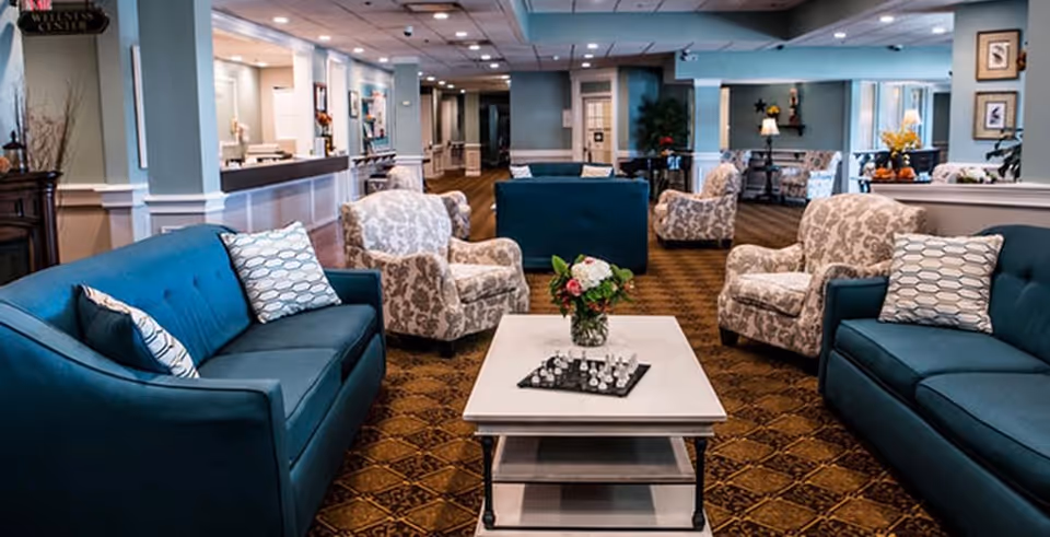 A spacious and well-lit lounge area in an assisted living community featuring two blue sofas and four patterned armchairs arranged around a white coffee table with a chess set and a vase of flowers. The room has a patterned carpet, light blue walls, and various decorative elements including lamps, framed pictures, and plants.