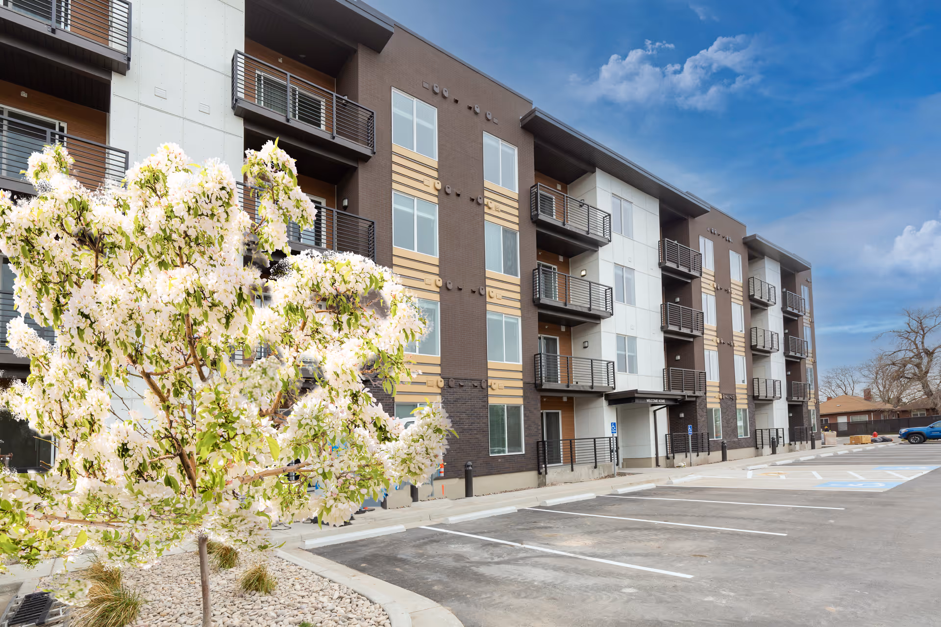 Exterior view of a modern multi-story senior living facility with balconies, a blooming tree in the foreground, and an empty parking lot under a partly cloudy sky.