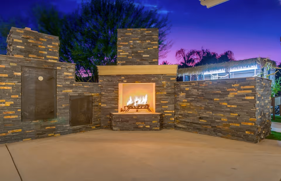 Outdoor stone fireplace and surrounding stone walls with a lit fire under a purple dusk sky.