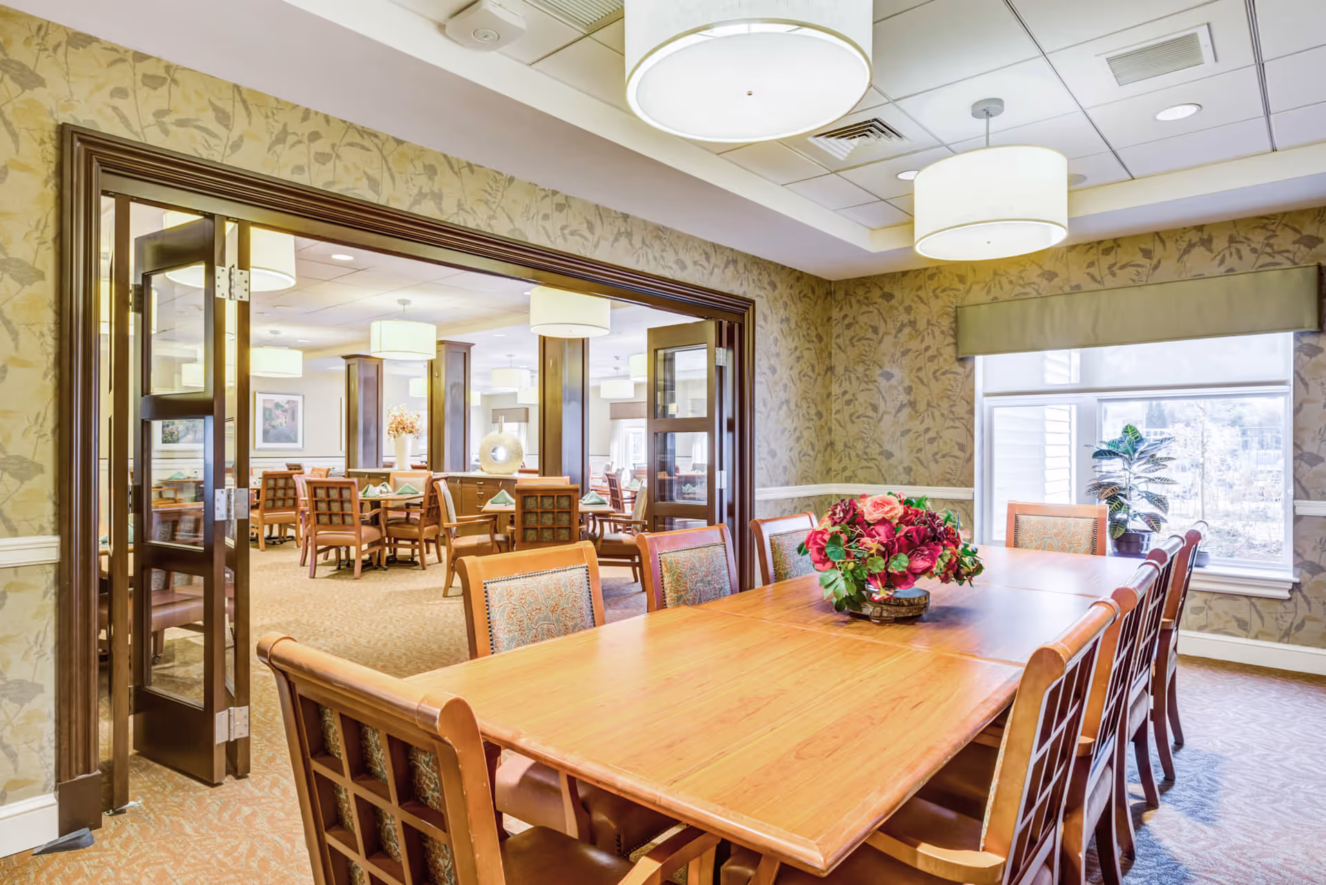 A bright dining room in a senior living facility featuring a long wooden table with chairs around it and a floral centerpiece. The room has patterned wallpaper, large windows with blinds, and modern ceiling lights. Through open double doors, another dining area with multiple tables and chairs is visible.