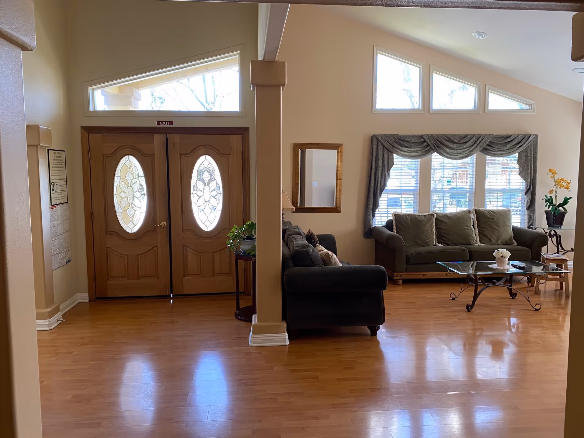 Interior view of a senior living facility showing a sitting area with two green sofas, a glass coffee table, a side table with a plant, and large windows with decorative curtains. To the left, there is a double wooden door with oval glass panels and an exit sign above it. The floor is wooden, and the walls are painted beige.