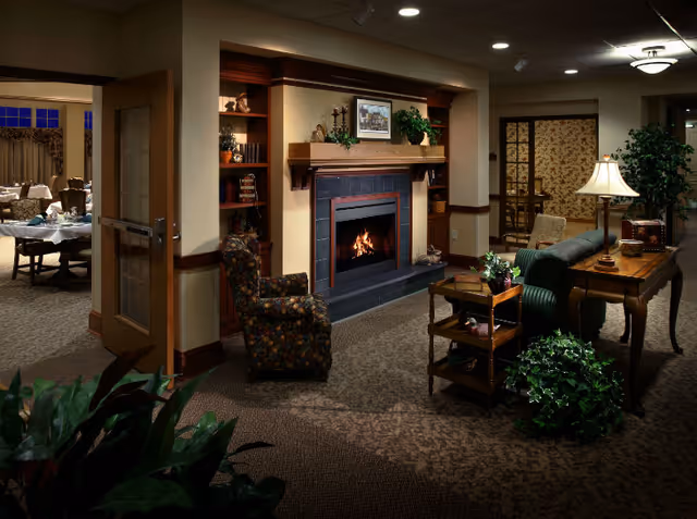 Cozy interior of a senior living facility featuring a lit fireplace with a mantel decorated with plants and framed pictures. There is a floral patterned armchair near the fireplace, a wooden table with a lamp and plants, and a carpeted floor. In the background, there is a dining area with tables and chairs visible through an open doorway.