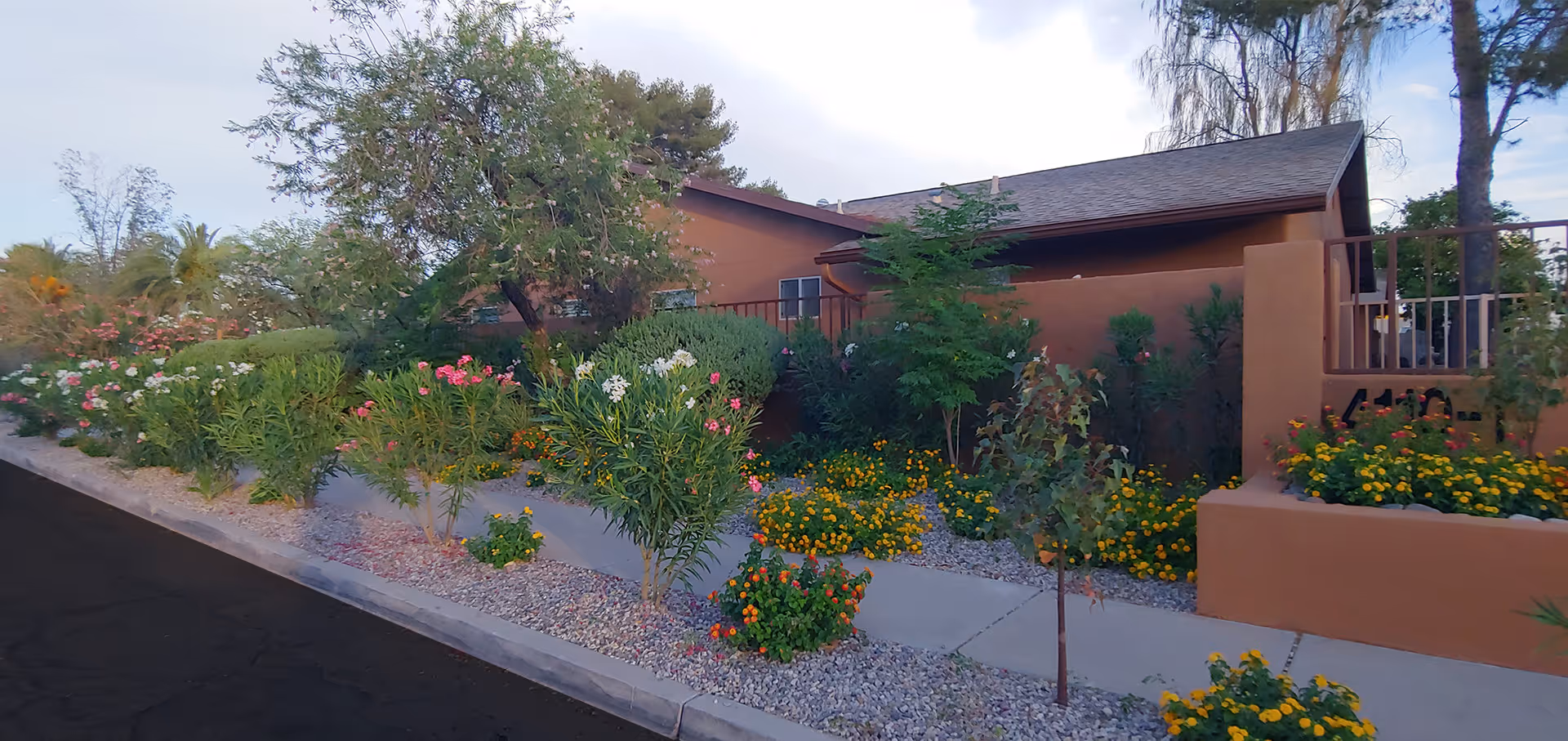 Exterior view of a single-story building with a brown facade, surrounded by a well-maintained garden featuring various flowering plants and shrubs along a sidewalk.