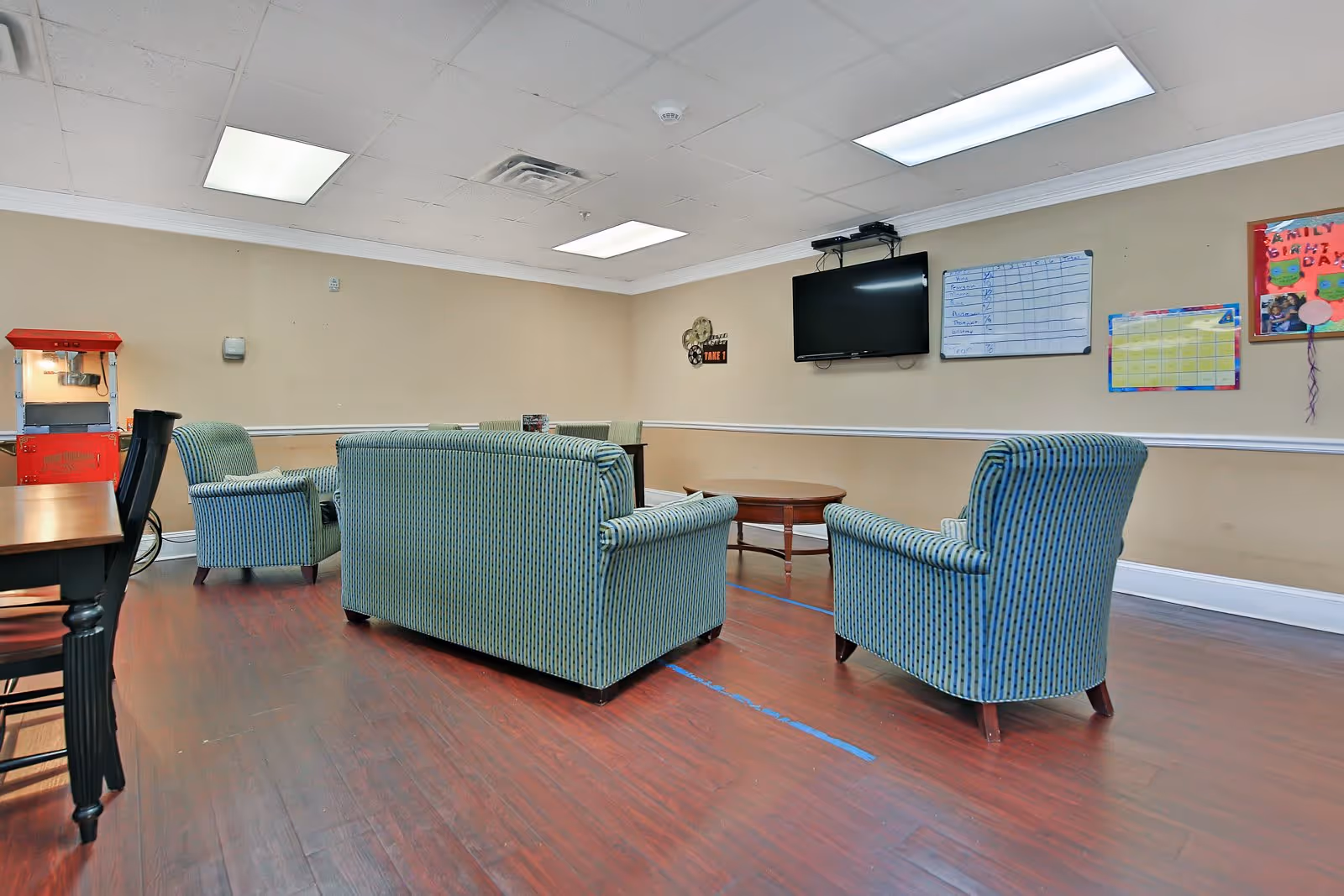 A common area in an assisted living facility with a striped blue sofa and two matching armchairs arranged around a wooden coffee table. A flat-screen TV is mounted on the beige wall above a whiteboard and colorful bulletin boards. There is a red popcorn machine in the corner and a wooden table with chairs on the left side. The room has wood flooring and fluorescent ceiling lights.