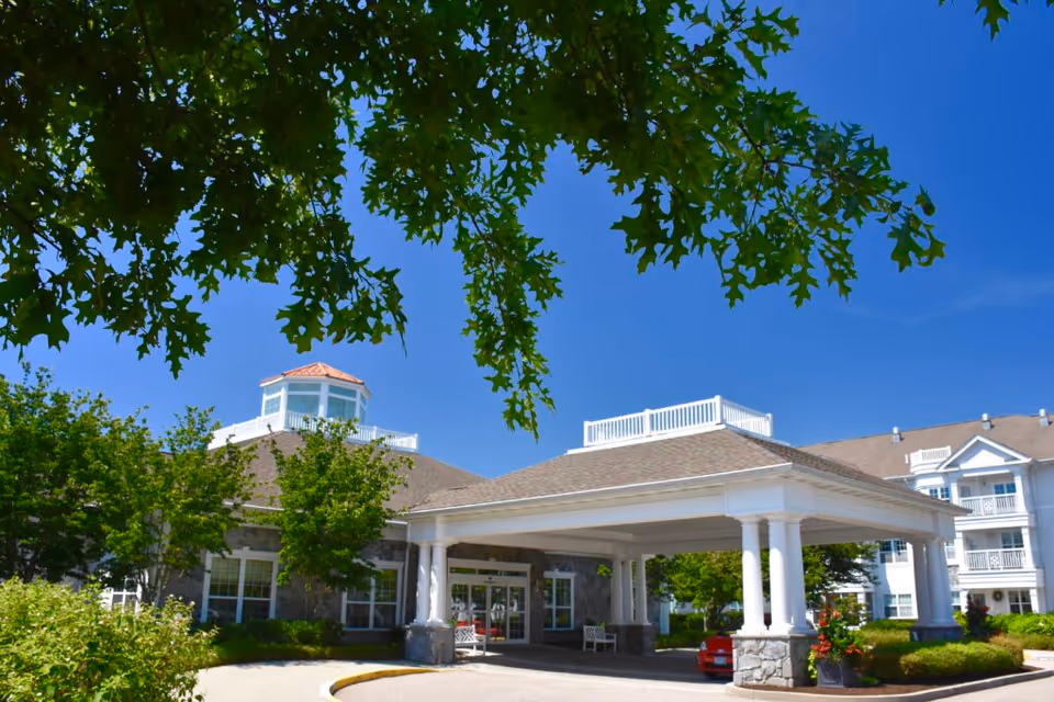 Exterior view of StoneRidge Senior Living facility showing the main entrance with a covered drop-off area supported by white columns, surrounded by green trees and bushes under a clear blue sky.