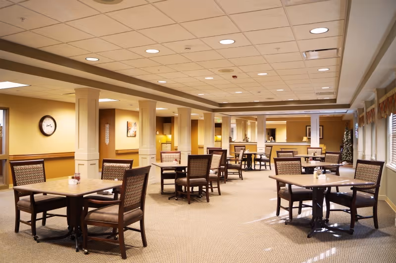 Spacious communal dining room with multiple tables and chairs, decorative columns, warm lighting, and windows along the right side.