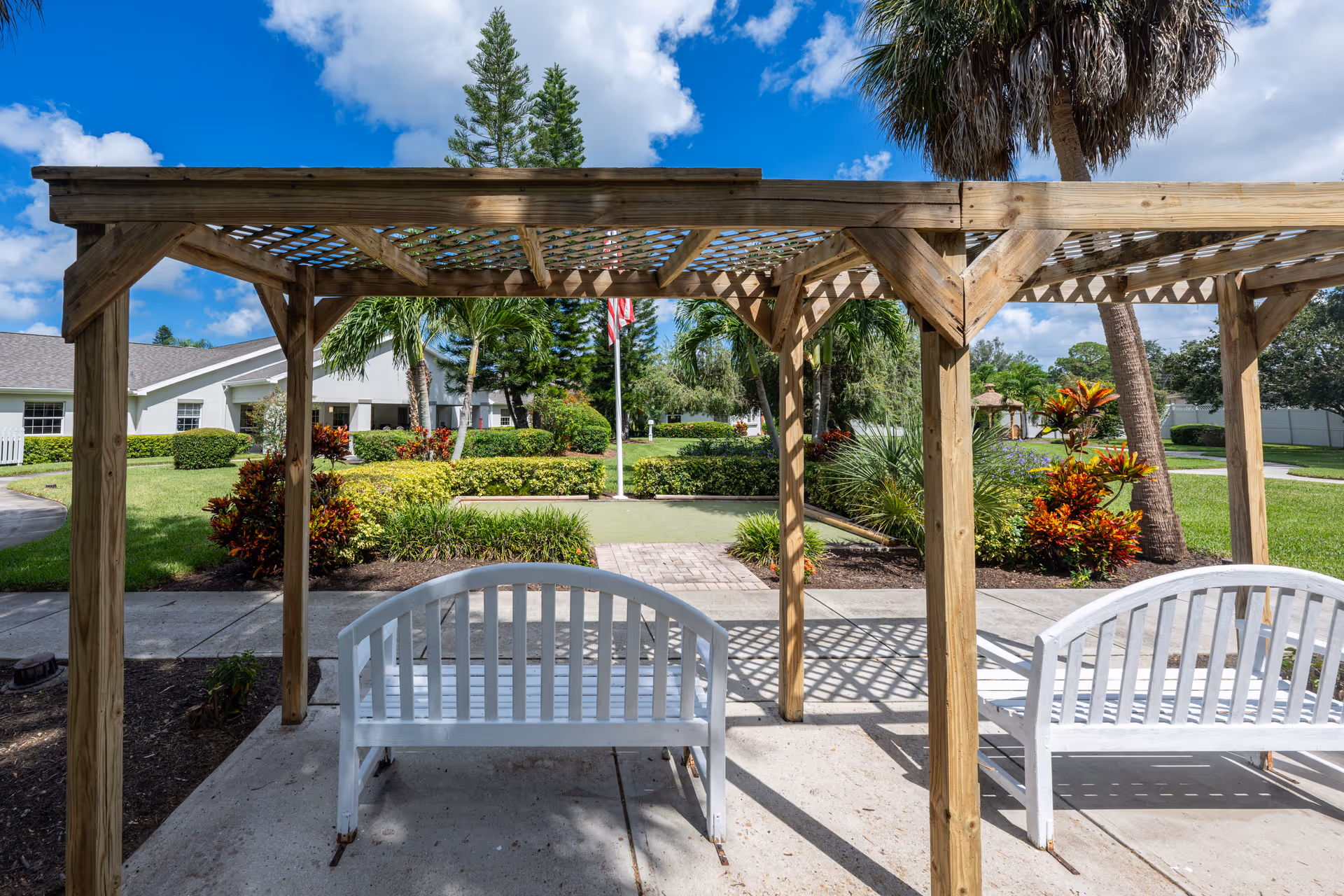 Outdoor seating area with two white benches under a wooden pergola. The area overlooks a well-maintained garden with various shrubs, palm trees, and an American flag in the background. A white building is visible on the left side under a partly cloudy blue sky.
