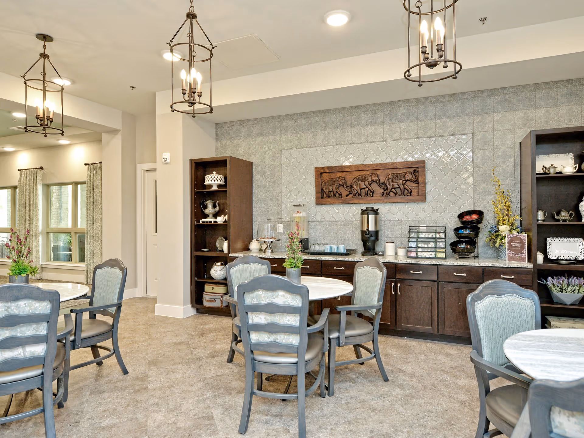A bright and elegant dining area with round tables and cushioned chairs. The back wall features dark wooden cabinets and shelves displaying decorative items and a beverage station with cups and dispensers. Above the cabinets is a decorative wooden panel with carved elephants. The room is lit by hanging lantern-style light fixtures and has large windows with patterned curtains.