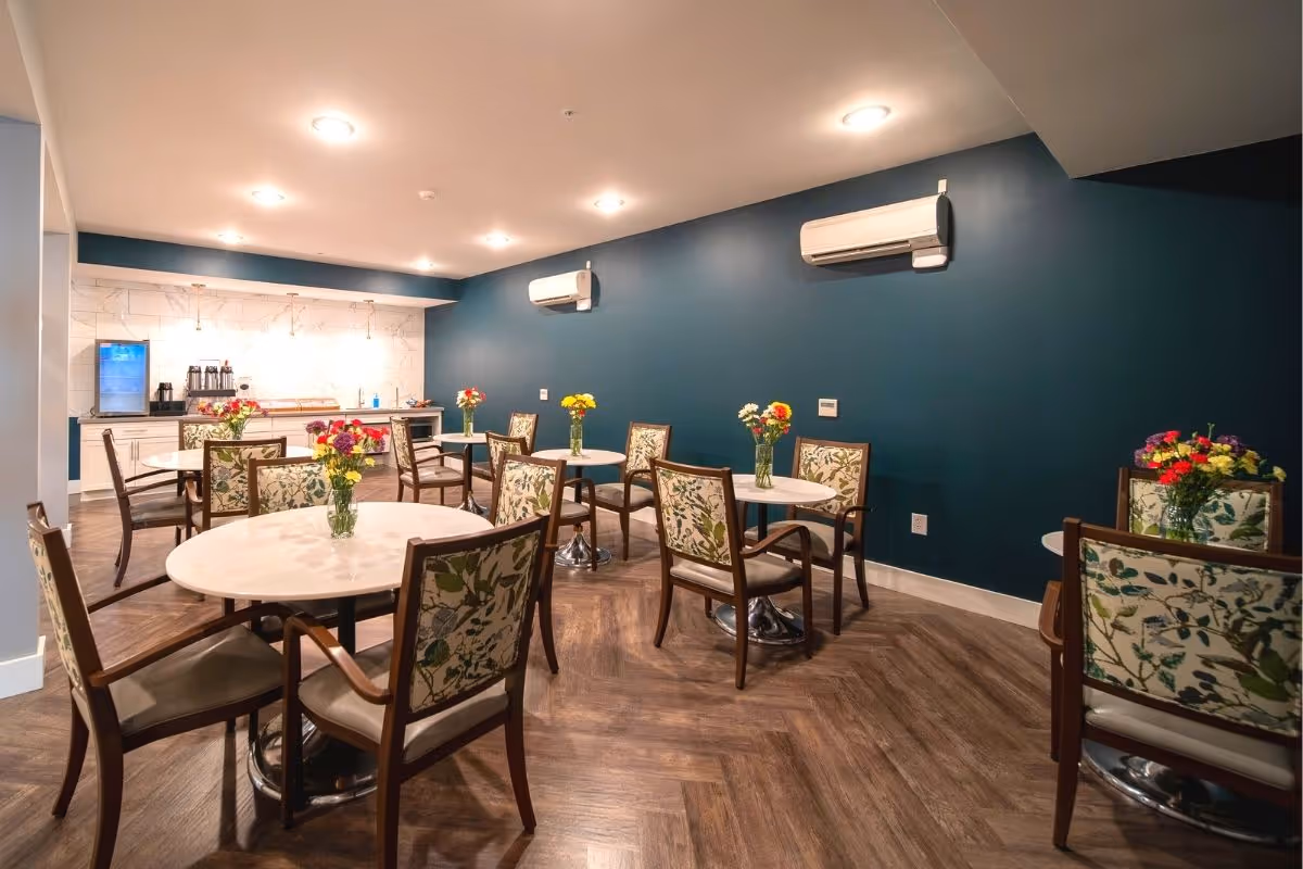 A dining room with several round tables, each surrounded by floral-patterned chairs. Each table has a vase with colorful flowers. The room has a dark teal wall with two air conditioning units mounted on it. The floor is wooden with a herringbone pattern. At the back of the room, there is a counter with a coffee machine, a small refrigerator, and some other items.