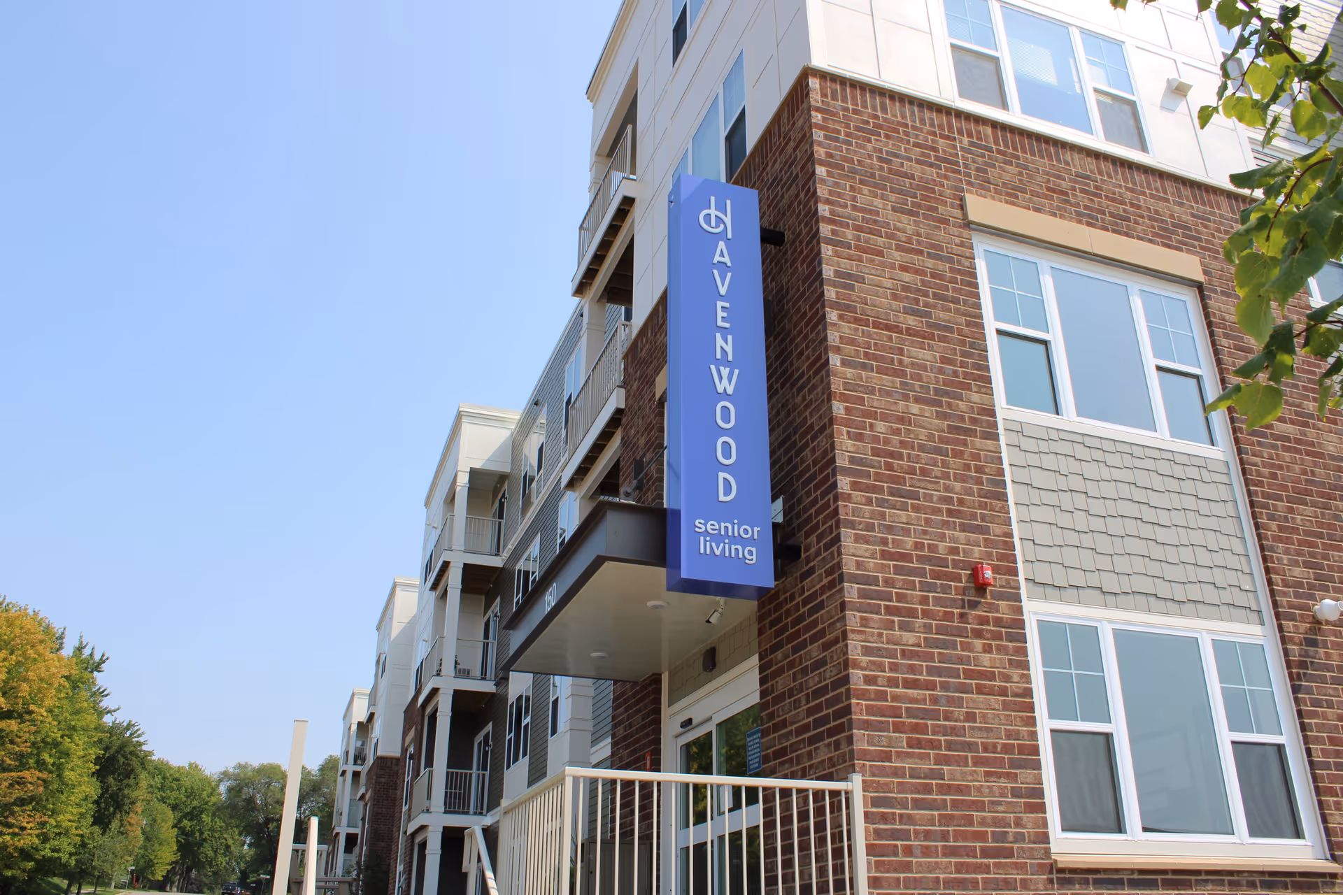 Exterior view of a multi-story senior living facility building with a brick and light-colored facade. A vertical blue sign on the building reads 'Havenwood senior living'. Trees and a clear blue sky are visible in the background.