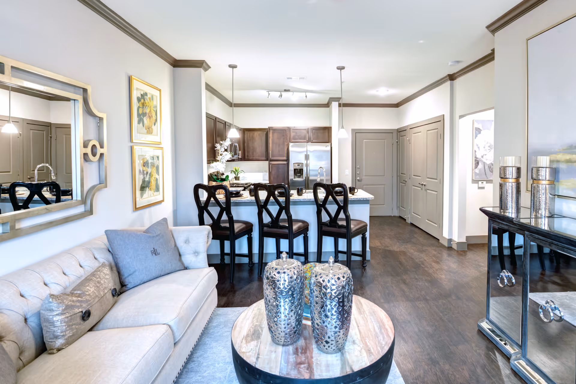 A modern open-concept living room and kitchen area with a beige tufted sofa adorned with pillows, a round wooden coffee table with two decorative silver vases, and a kitchen island with four dark wooden bar stools. The kitchen features dark wood cabinets, stainless steel refrigerator, and pendant lighting. The walls are light-colored with crown molding, and there is a large mirror and framed floral artwork on the walls.