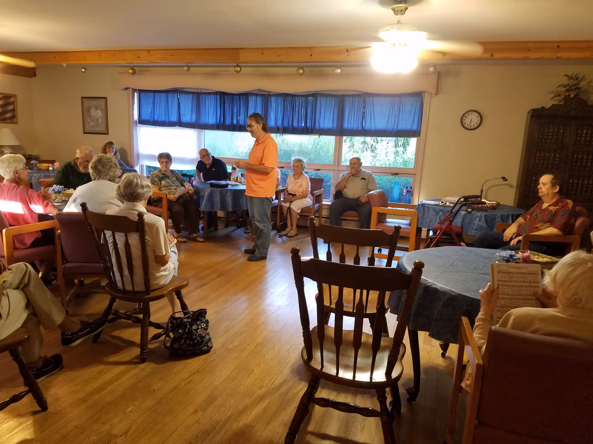 A group of elderly people seated in a common room with wooden floors and blue tablecloth-covered tables. One man in an orange shirt stands in the middle speaking to the group. The room has large windows with blue curtains, a ceiling fan, and wooden furniture.