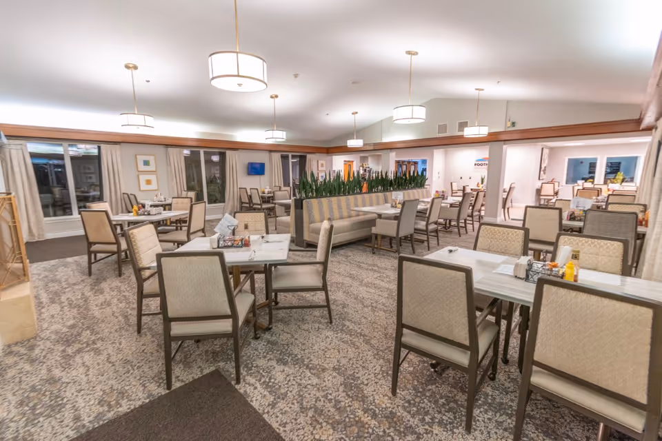 A spacious dining room with multiple tables and chairs arranged neatly. The room features carpeted flooring, large windows with curtains, modern pendant lights hanging from the ceiling, and a central seating area with a cushioned bench and plants as a divider.