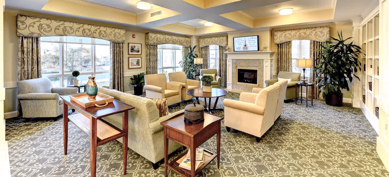 A spacious and well-lit living room with multiple beige armchairs and sofas arranged around a round wooden coffee table. The room features large windows with patterned curtains, a fireplace with a TV mounted above it, decorative plants, and side tables with lamps. The carpet has a patterned design, and the walls are painted in warm tones.