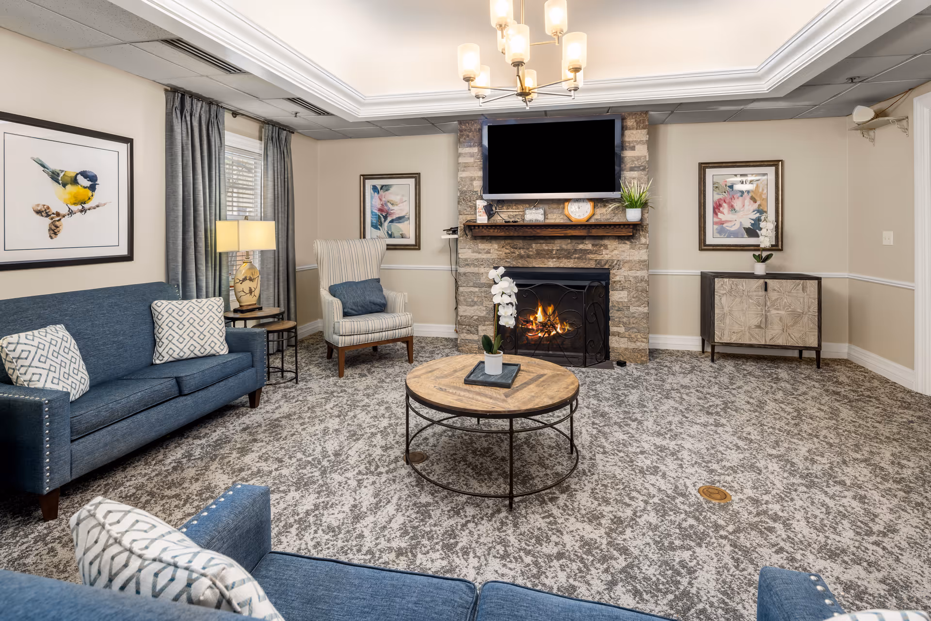 A cozy living room with a stone fireplace and a lit fire beneath a mounted flat-screen TV. The room features a blue sofa and armchair with patterned cushions, a round wooden coffee table with a small potted plant, framed floral and bird artwork on the walls, a table lamp, and a sideboard with a potted orchid. The carpet is patterned in gray tones, and the ceiling has a modern chandelier.