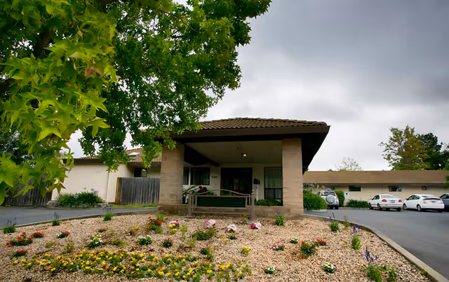 Covered entrance/porte-cochere of a single-story building with a landscaped rock and flower bed and parked cars nearby under a cloudy sky.