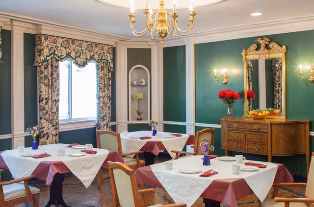 A dining room with four tables covered in white and burgundy tablecloths, each set with plates, cups, and napkins. The room features green walls with white trim, a window with floral curtains, a wooden sideboard with a large ornate mirror above it, and a chandelier hanging from the ceiling. Small flower arrangements are placed on each table and on the sideboard.